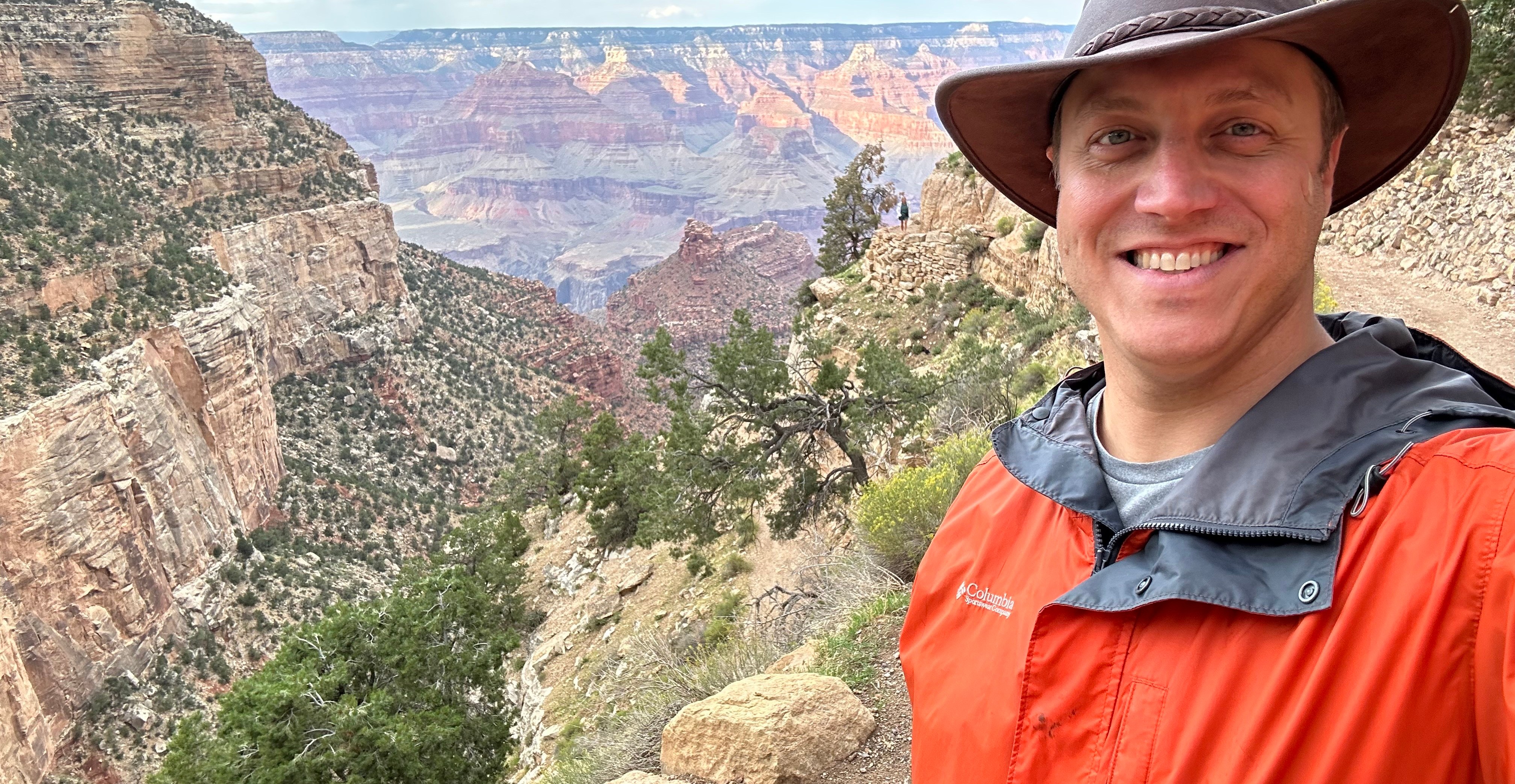 A man in a leather hat and an orange raincoat stands on the edge of a canyon.