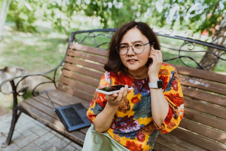 A woman on a park bench talking on the phone with an earbud in her ear.