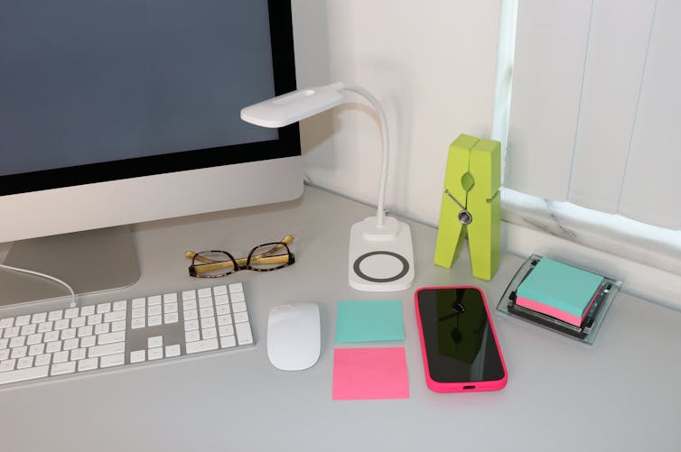 A computer, smartphone and mouse sit on a desk next to a lamp and some notepapers.