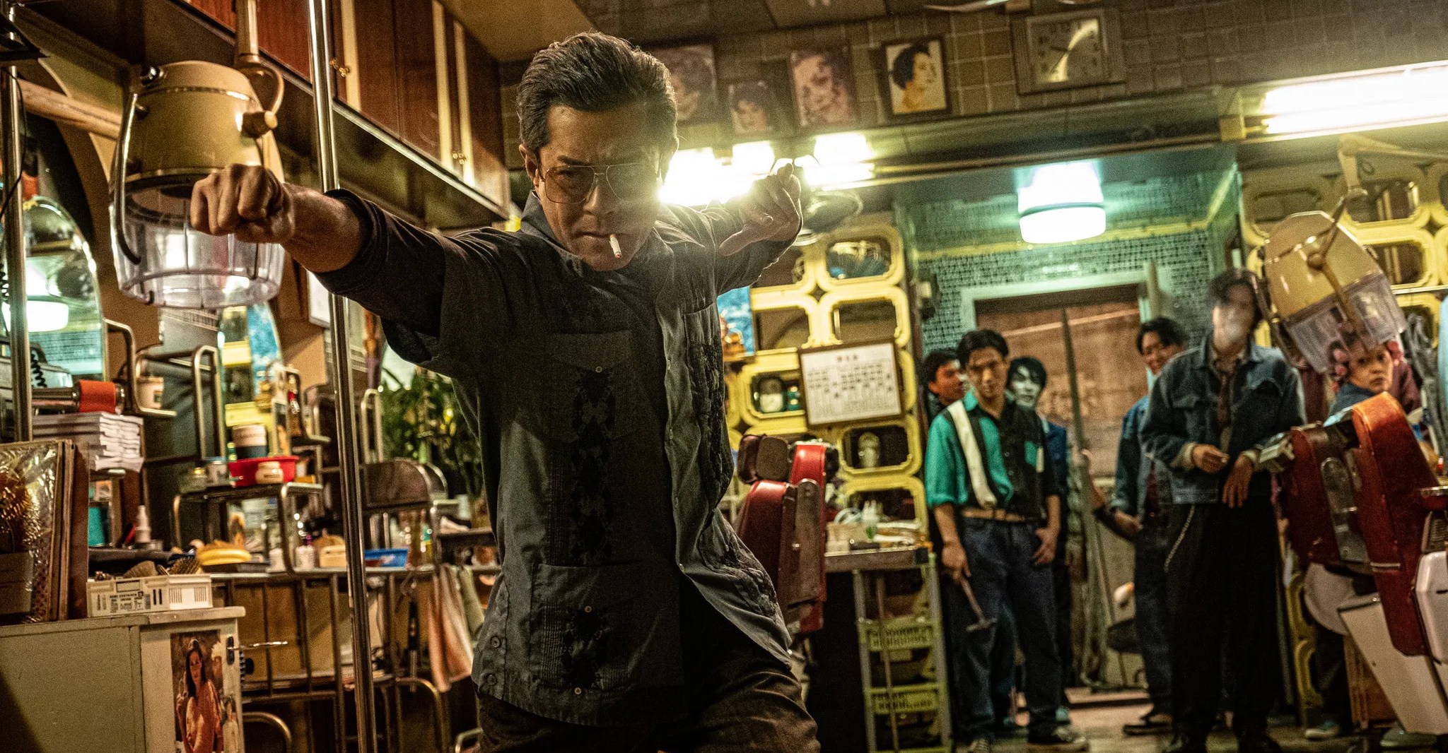 A man wearing glasses and smoking a cigarette holding a pose with a group of men standing in the background in Twilight of the Warriors: Walled in.