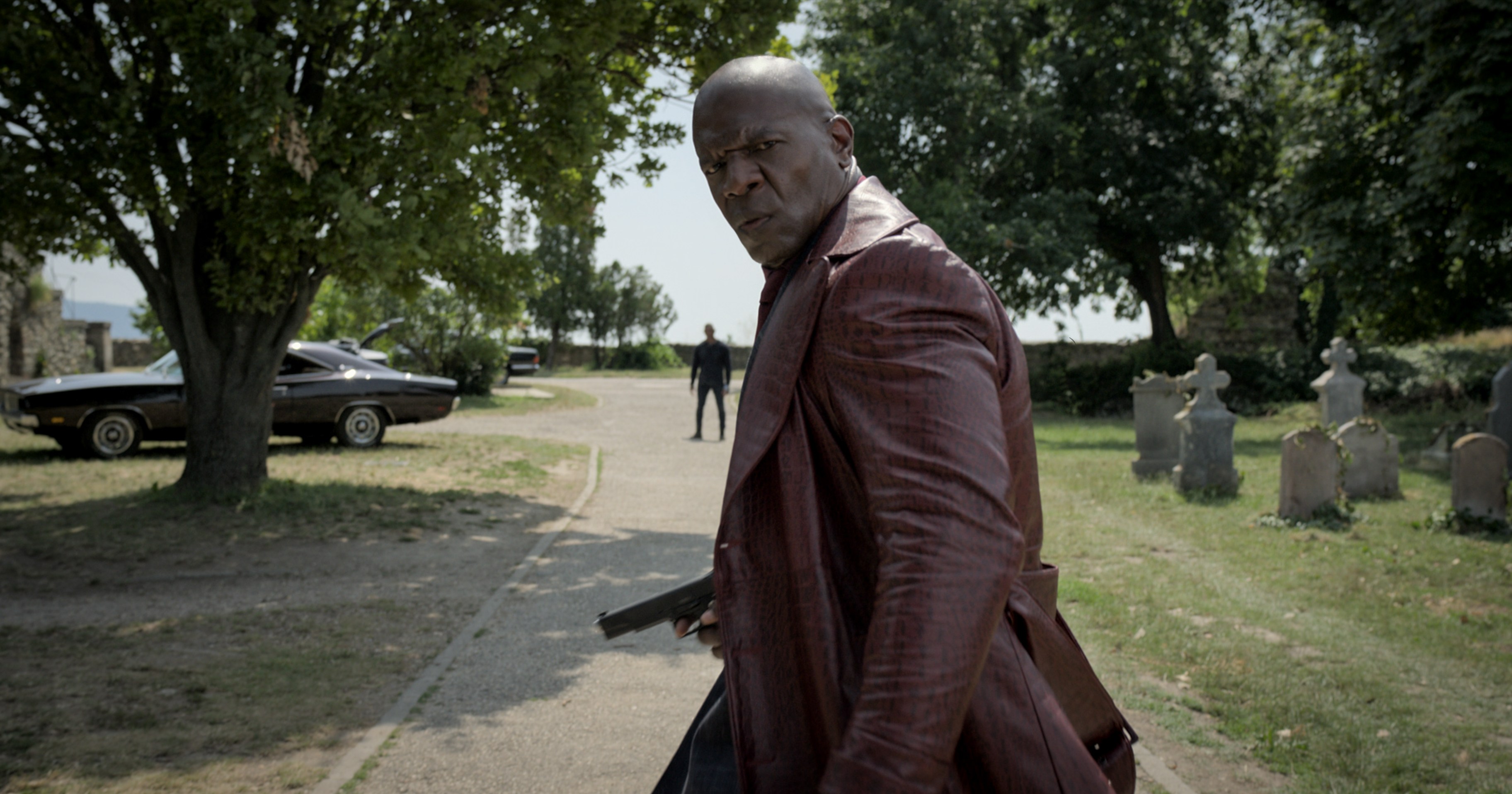 Terry Crews in The Killer’s Game, wearing a long leather coat and holding a gun while looking at the camera, confused