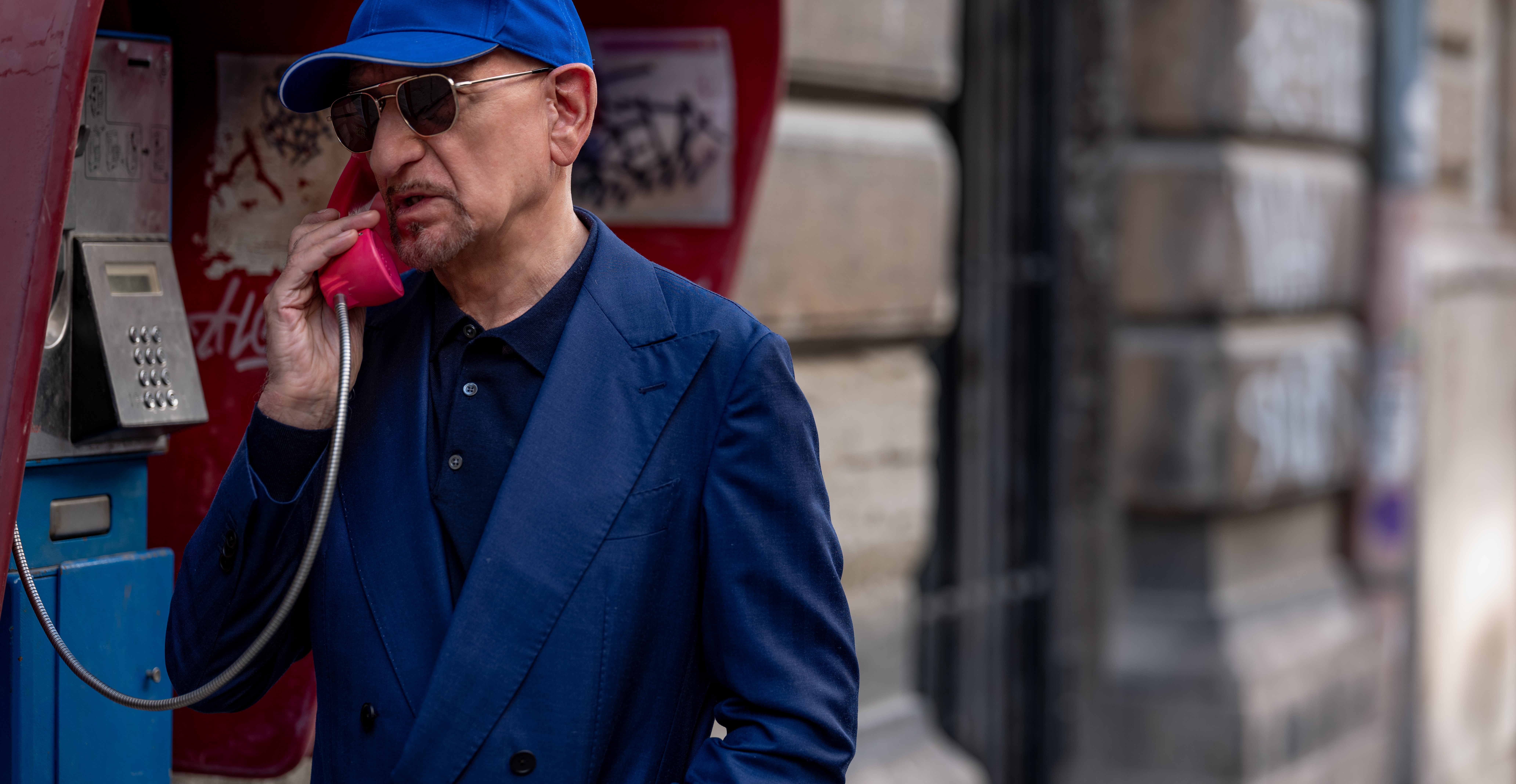 Ben Kingsley, wearing a blue hat and sunglasses, talking on a phone in  a phone booth in The Killer’s Game