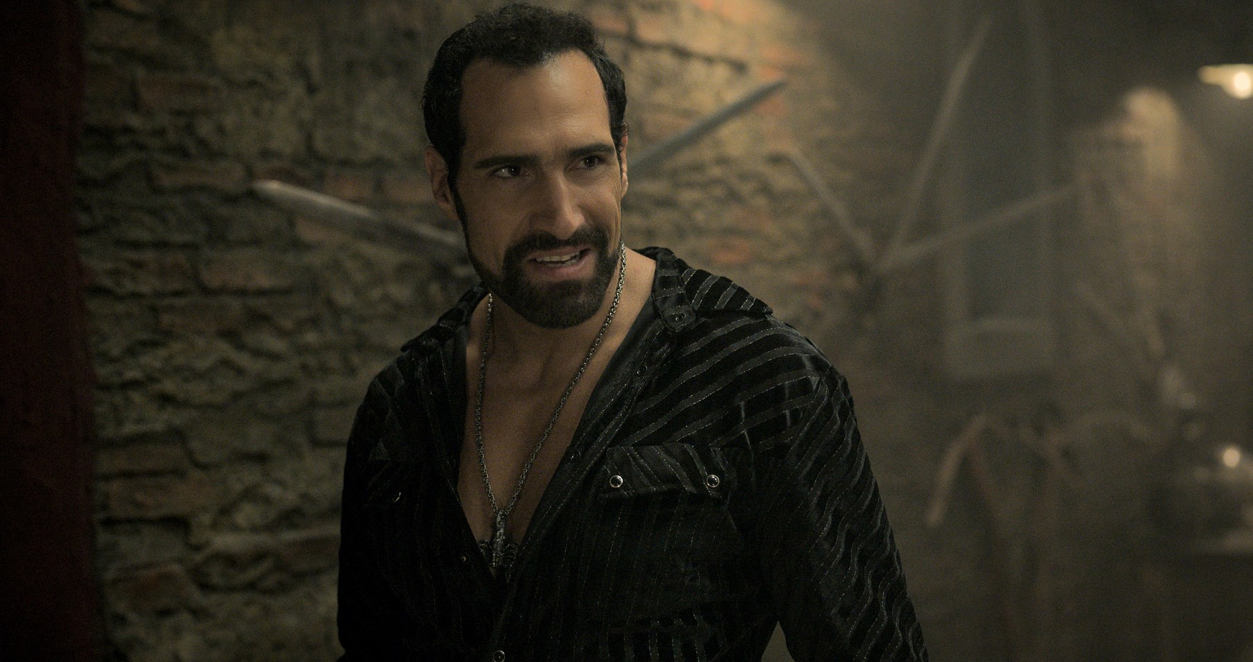 Marko Zaror as Botas in The Killer’s Game, smiling, with his shirt open to show his long necklace