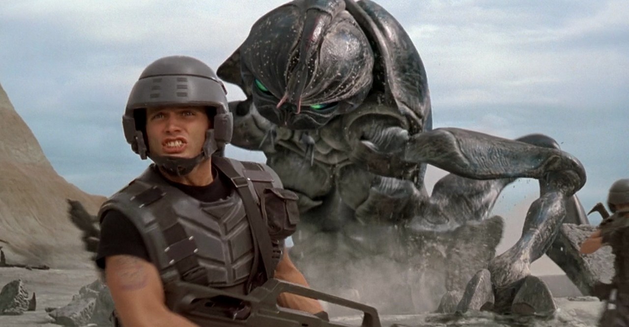 A man in soldier armor standing in front of a gigantic insect in Starship Troopers.