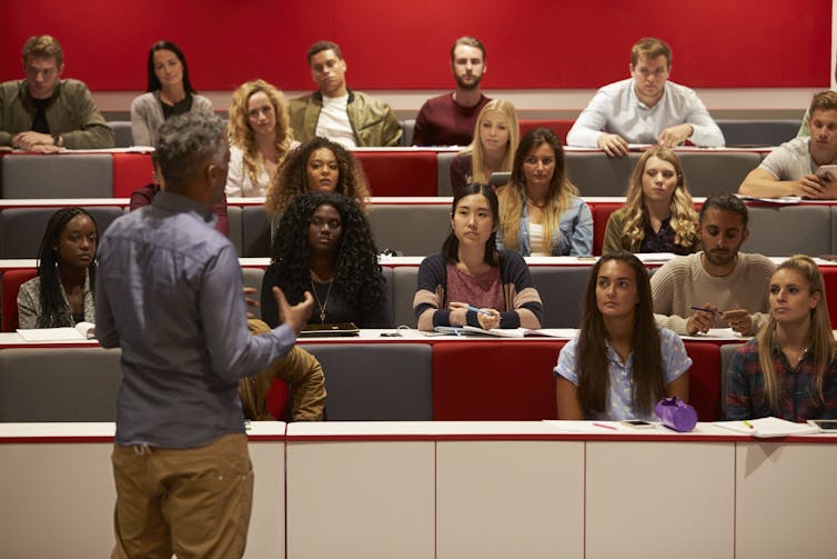 A man stands in front of a group of young people who are in tiered seating in a lecture hall.