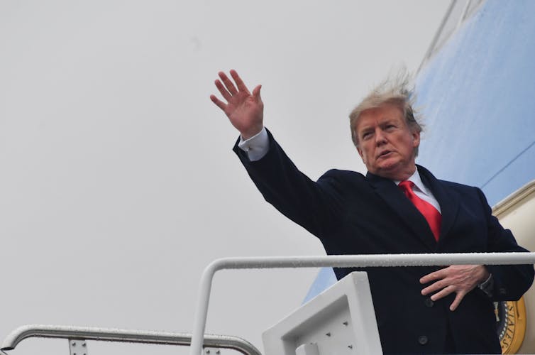 a man in a business suit waves from in front of the door to an airplane