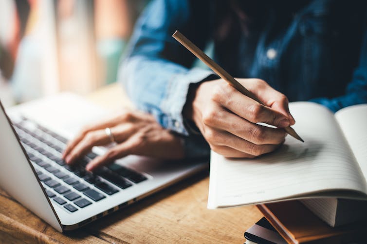 One hand types on a computer, while another writes in a notebook.