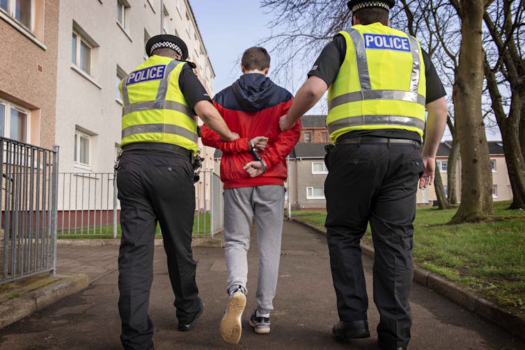 Staged stock photo, view from behind of a young boy in handcuffs being escorted by two police officers