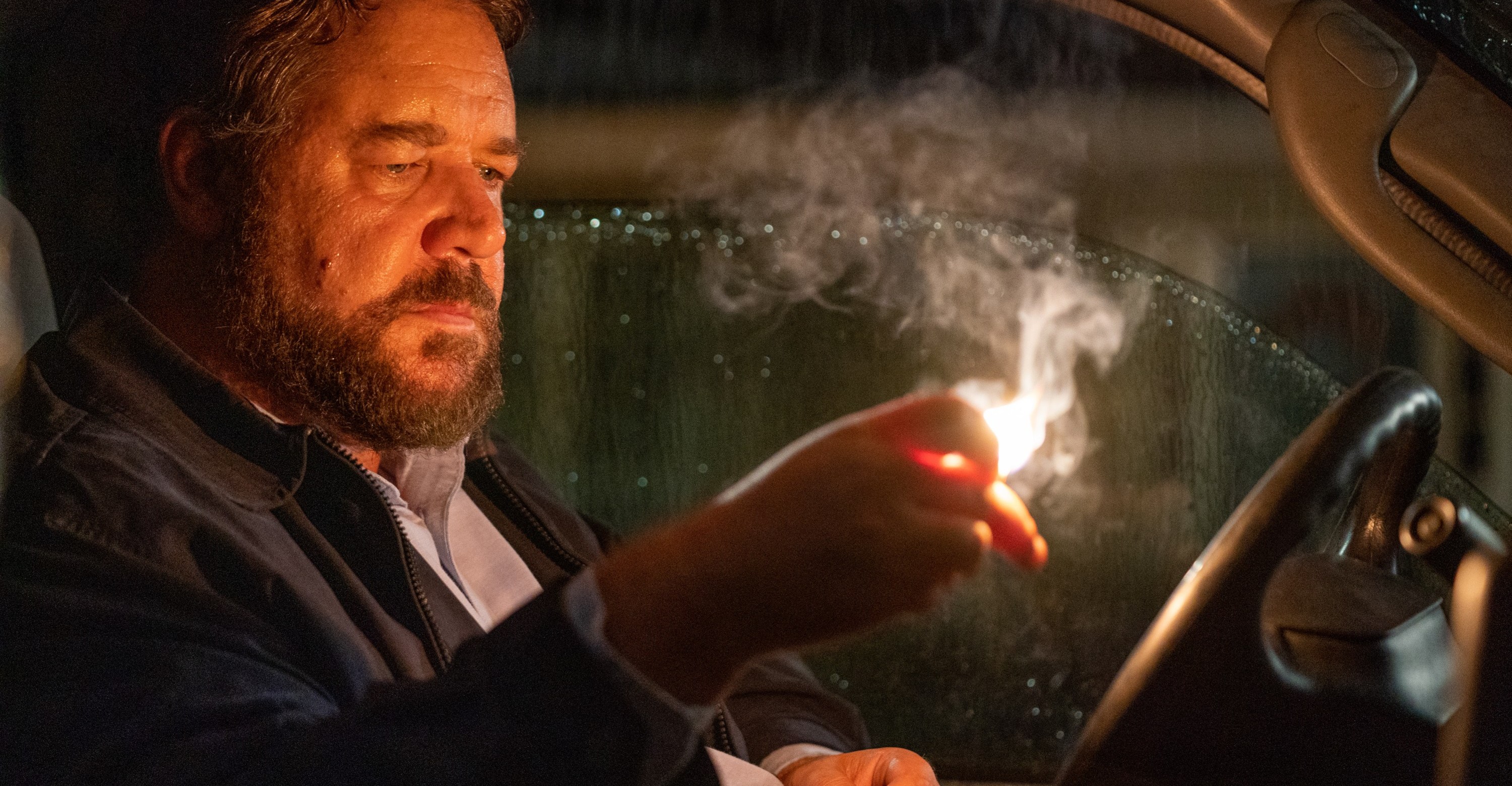 Russell Crowe sitting in the front of a pickup truck in Unhinged looking at a lit match in his hand