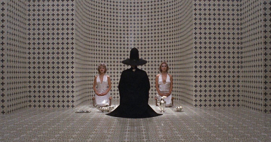 A person in a black cloak and hat kneeling in the middle of a checkered patterned room flanked by women in white outfits in The Holy Mountain.