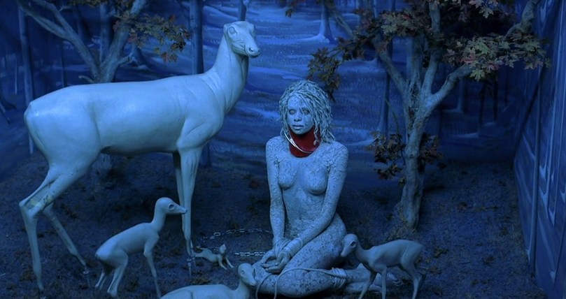 A woman covered in white makeup and bound in a menagerie surrounded by statues of deer and fawn in The Cell (2000).