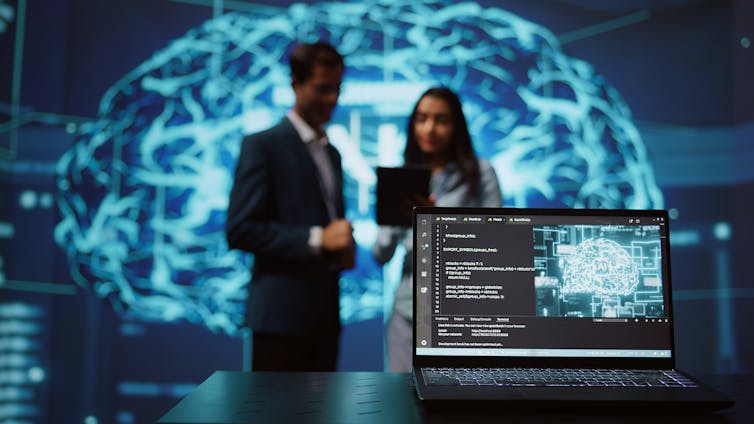 An open laptop with an image of a brain and the word AI is seen in the foreground while the blurred image of two people have a conversation in front of a holographic brain in the background