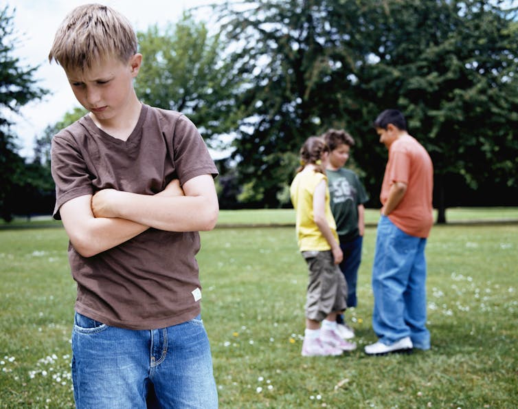 dejected tween boy stands with arms crossed in foreground, in background three other boys talk together